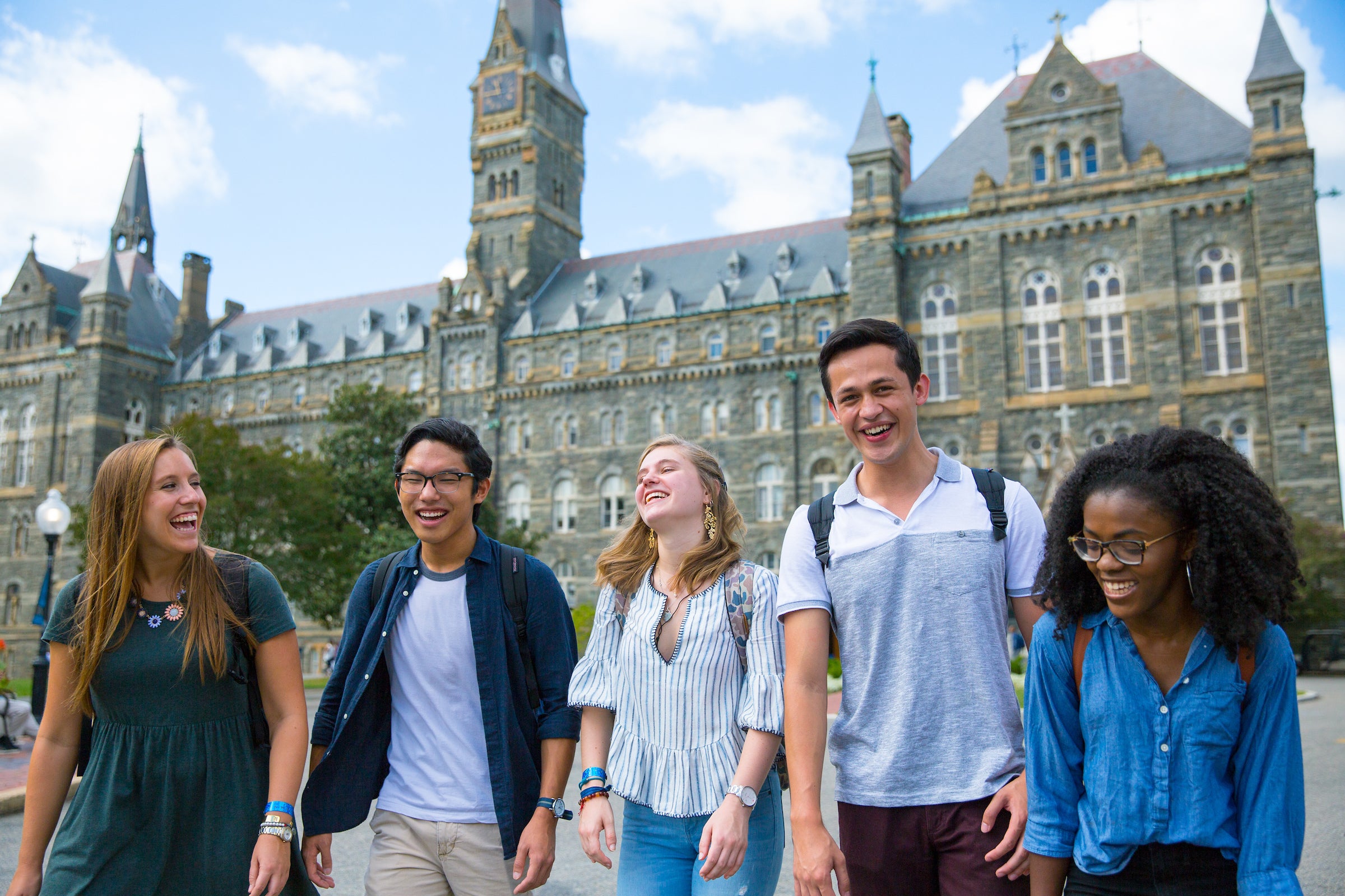 a group of five students walk shoulder to shoulder, smiling, with Healy Hall in the background