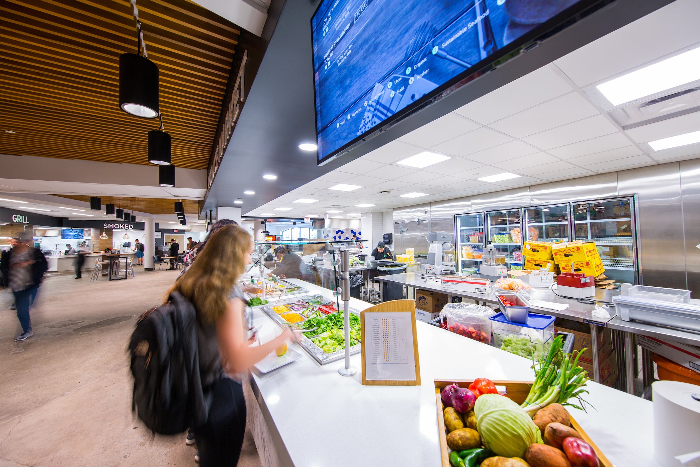 student selecting a meal at the cafeteria, which is brightly lit, shows menus on TVs above, and has lots of colorful fruits and vegetables
