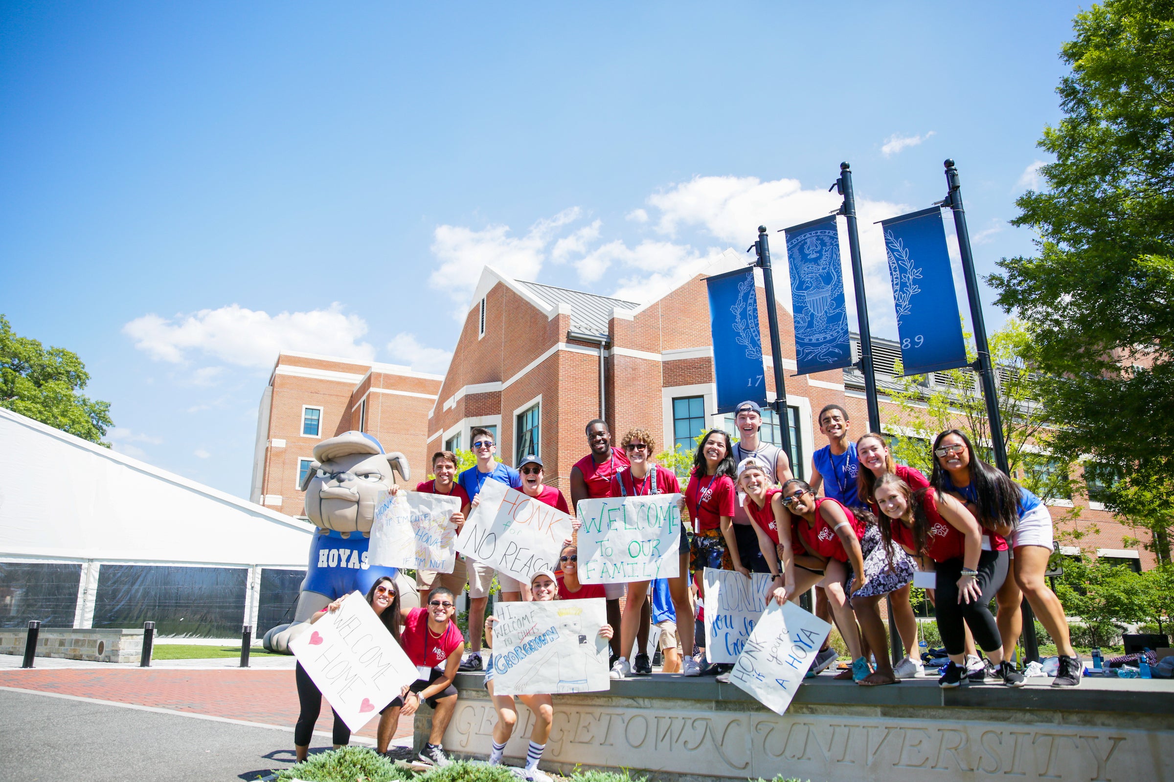 a group of 10-15 students in red t shirts welcomes freshman to campus with signs